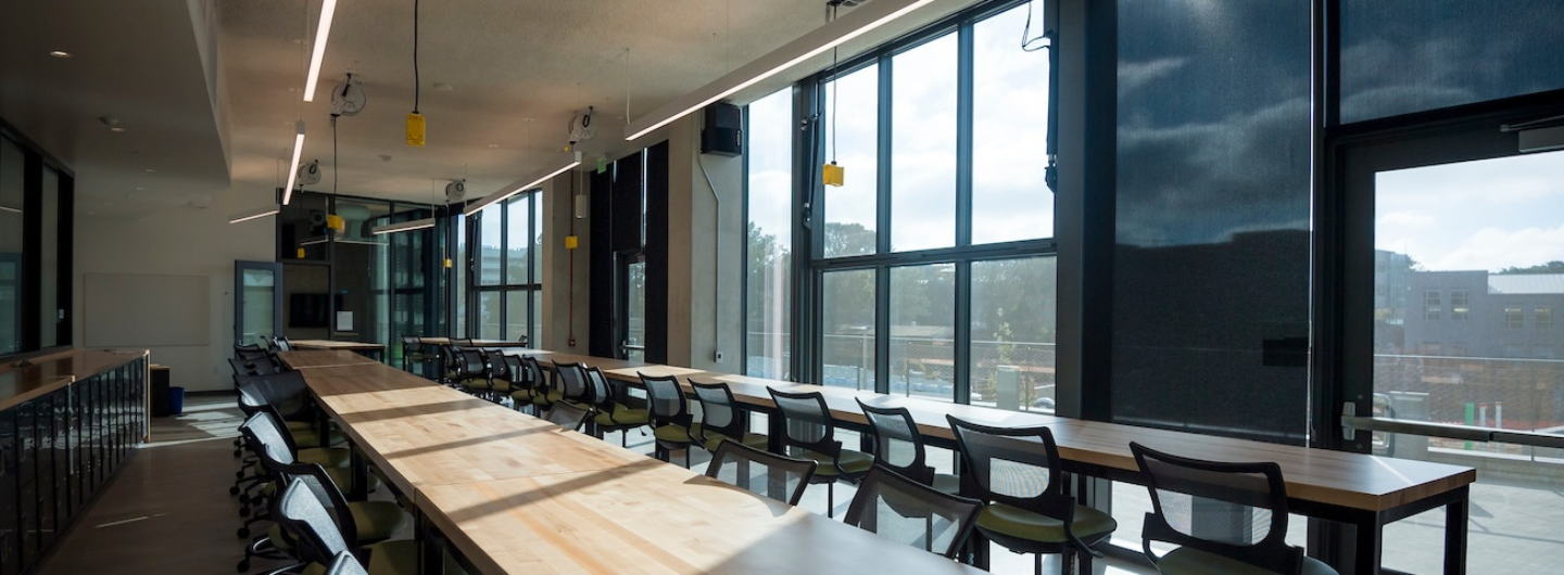 A large, empty room with long wooden tables and black chairs, featuring floor-to-ceiling windows along one side, offering a view of the outside